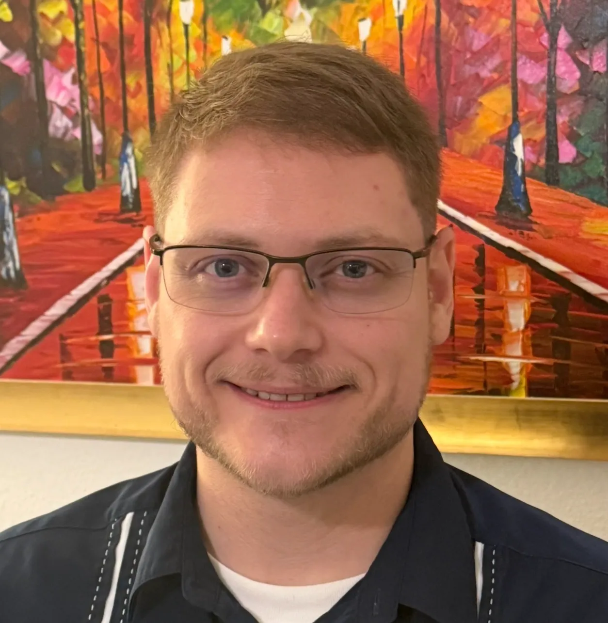 Aaron, smiling, wearing glasses and a dark button-up shirt, standing in front of a colorful painting of an autumn park path.