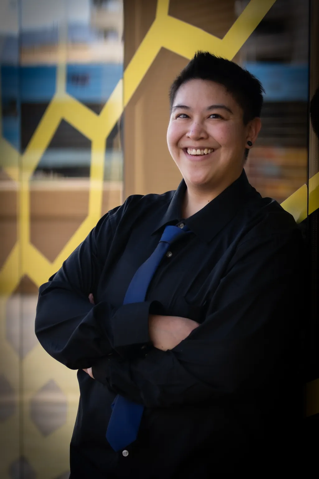 Jamie, smiling with arms crossed, wearing a dark shirt and blue tie, standing in front of a yellow geometric pattern.